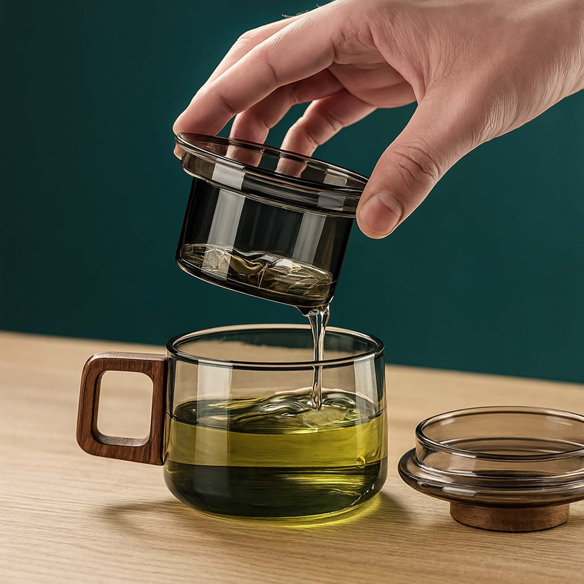 Hand pouring liquid from a glass container into a glass mug with wooden handle on a wooden surface.