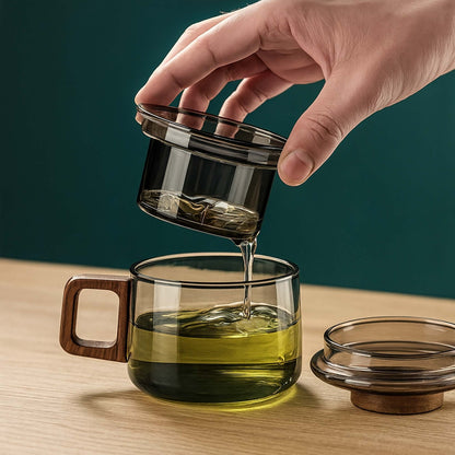 Hand pouring liquid from a glass container into a glass mug with wooden handle on a wooden surface.