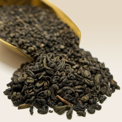 Dry black Gunpowder tea leaves in a golden scoop on a light background